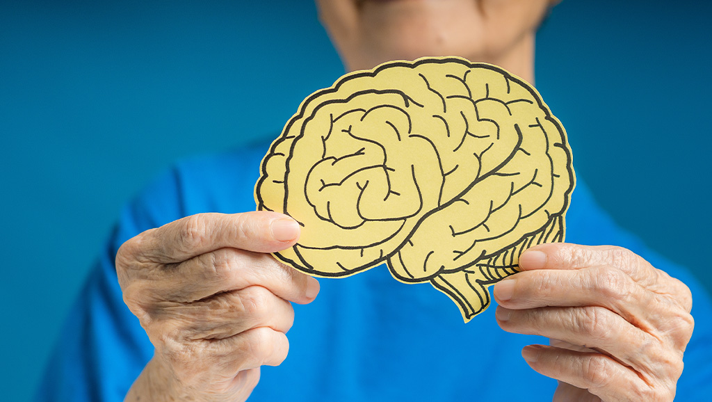 Hands of an older woman holding a brain shape made from yellow paper against a blue background.
