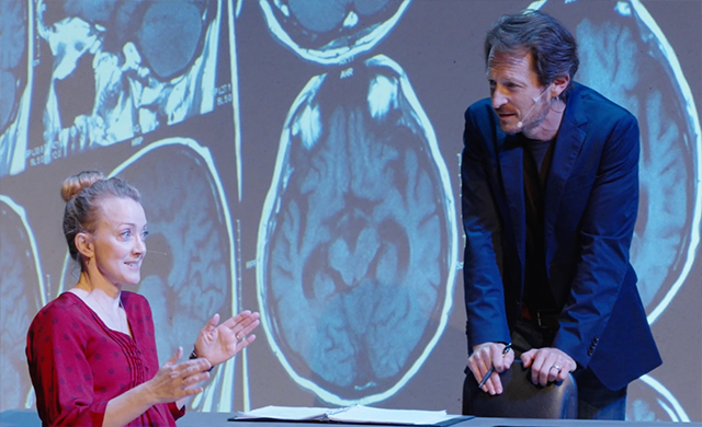 Photo of two actors on stage with brain imaging in the background